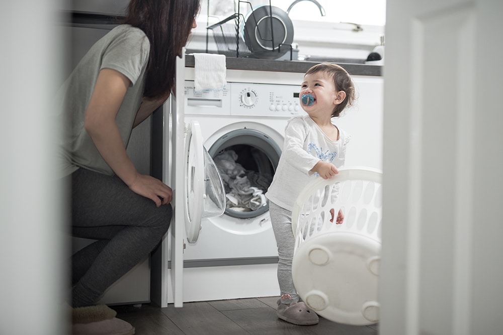 An Asian mother and her daughter enjoy and laugh doing the laundry in the kitchen of a house in Edinburgh, Scotland, United Kingdom, where a washing machine can be seen on the background